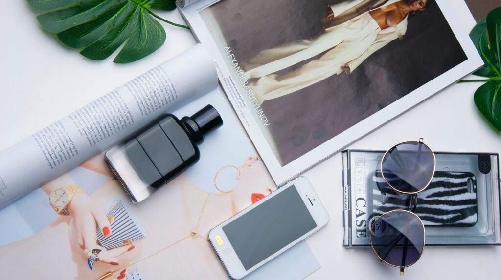 Flat lay of fashion items including sunglasses, perfume, and a magazine on a white surface.